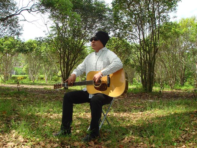 写真：木立の中で堀内佳さんが椅子に座ってギターを持っています